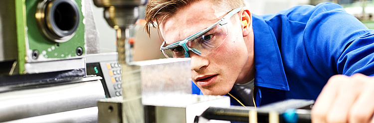 Ein Mann in einem blauen Arbeitskittel und Schutzbrille konzentriert sich auf die Prüfung oder Einstellung einer Maschine in einer Fertigungsumgebung.