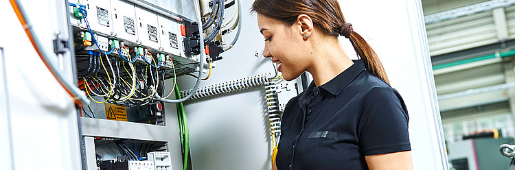 Eine Frau in Arbeitskleidung überprüft die Verdrahtung in einem elektrischen Schaltschrank in einer industriellen Umgebung.