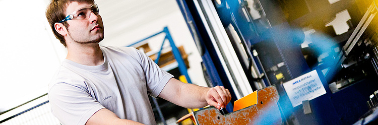 Ein Mann in einem hellen T-Shirt und Schutzbrille bedient oder überprüft eine industrielle Maschine.