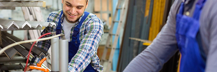 Mann im karierten Hemd und Latzhose arbeitet lächelnd an Maschinenteilen in einer Werkstatt.