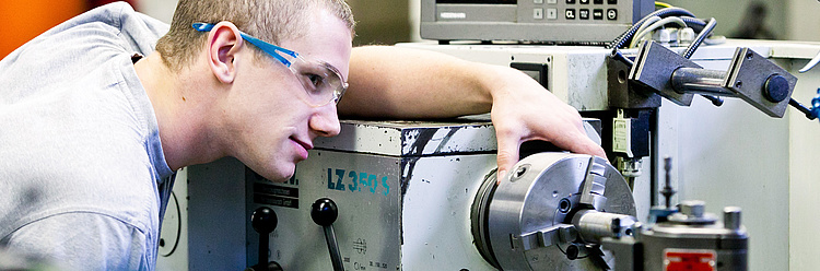 Ein junger Mann mit kurzen Haaren und Schutzbrille beugt sich konzentriert über eine große Drehmaschine, um an einem metallischen Werkstück zu arbeiten, in einer industriellen Werkstatt