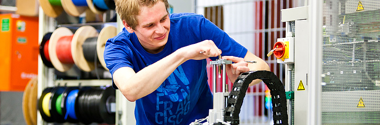 Ein junger Mann in einem blauen Shirt arbeitet an einer technischen Anlage mit Kabelschlauch und Werkzeug.
