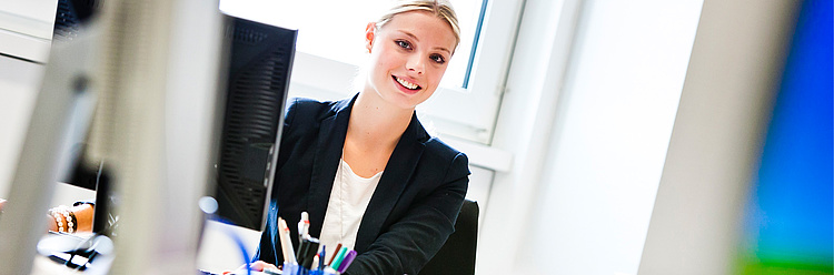 Eine junge Frau in Business-Kleidung arbeitet an einem Schreibtisch vor einem Computermonitor und lächelt.