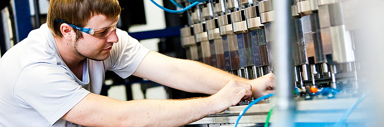 Ein Mann in einem Labor oder Werkstattumfeld arbeitet an einer komplexen Maschine oder Ausrüstung. Er trägt Schutzbrille und konzentriert sich auf seine Arbeit.