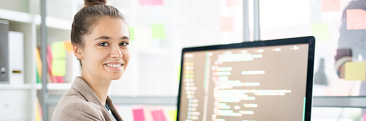 Junge Frau arbeitet lächelnd an einem Computer mit Programmiercode auf dem Bildschirm in einem hellen Büro.