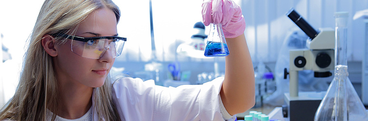 Eine Wissenschaftlerin im weißen Laborkittel und mit Schutzbrille betrachtet eine Flasche mit blauer Flüssigkeit in einem Labor.