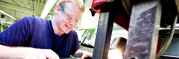 Ein lächelnder Mann in einem dunkelblauen Shirt arbeitet an einer Maschine mit leuchtenden Funken, wahrscheinlich beim Schweißen oder Schleifen.
