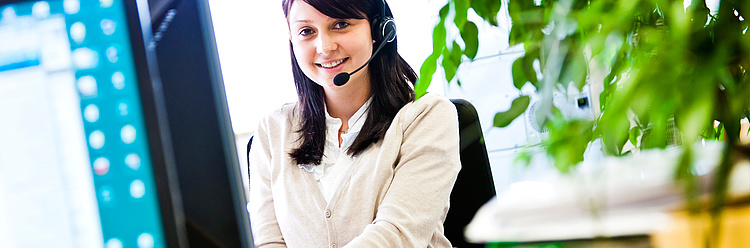 Eine Frau mit einem Headset sitzt vor einem Computerbildschirm und lächelt, während sie vermutlich in einem Callcenter oder Kundens