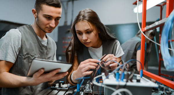 Informationselektronikerin mit Schutzhelm und Warnweste blickt vor einer Industrieanlage konzentriert auf ein Notebook.