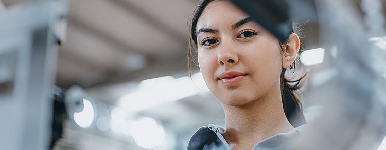 Eine junge Frau blickt selbstbewusst in die Kamera, während sie in einer industriellen Umgebung arbeitet. Unscharfe Maschinen und helle Beleuchtung sind im Hintergrund zu erkennen.