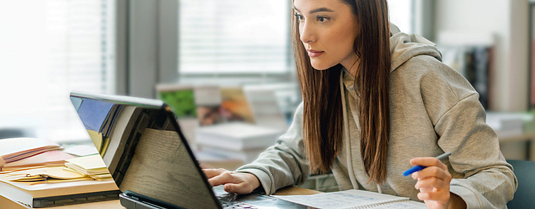 Eine junge Frau in einem hellgrauen Hoodie sitzt konzentriert an einem Schreibtisch, arbeitet an einem Laptop und hält einen Stift in der Hand. 