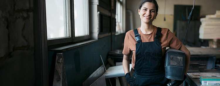 Eine lächelnde Frau in Arbeitskleidung mit Schweißerhelm in der Hand steht in einer Werkstatt. Im Hintergrund sind Werkzeuge und Arbeitsmaterialien zu erkennen.