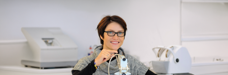 Eine Frau mit Brille und einem grauen Strickjacke sitzt in einem Labor und justiert ein optisches Gerät.