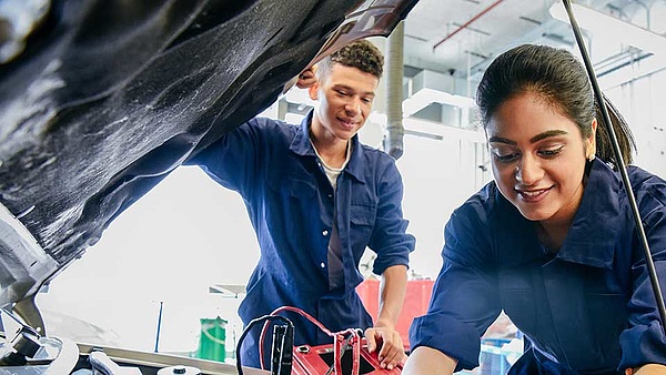 An einer offenen Motorhaube arbeiten zwei Auszubildende am Motorblock