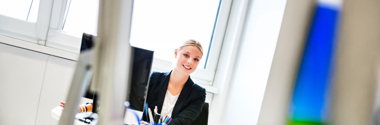 Eine junge Frau in Business-Kleidung arbeitet an einem Schreibtisch vor einem Computermonitor und lächelt.