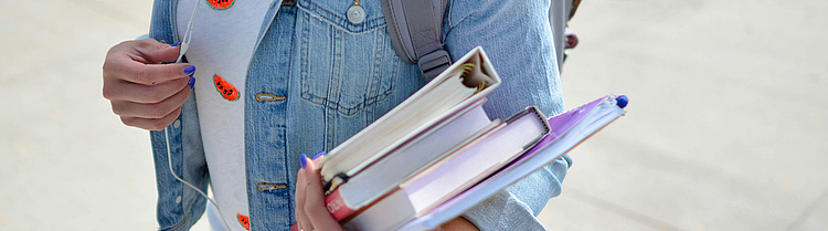 Eine Person in einer Jeansjacke trägt mehrere Bücher und einen grauen Rucksack. 