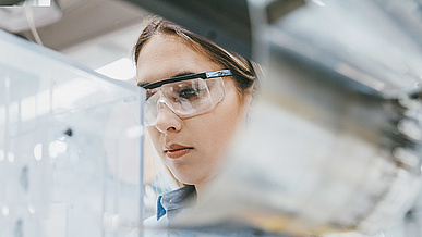 Eine junge Frau mit Schutzbrille arbeitet konzentriert an einer Maschine oder in einem Laborumfeld. 