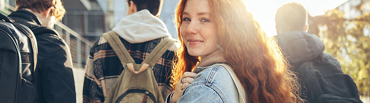 Junge Frau mit roten Locken und Jeansjacke blickt lächelnd über die Schulter, während sie mit einer Stofftasche und anderen Studierenden unterwegs ist.