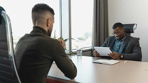 Ein Mann in einem grünen Blazer sitzt in einem hellen Büro und hört einem anderen Mann in einem grauen Anzug zu, der ein Dokument überprüft.