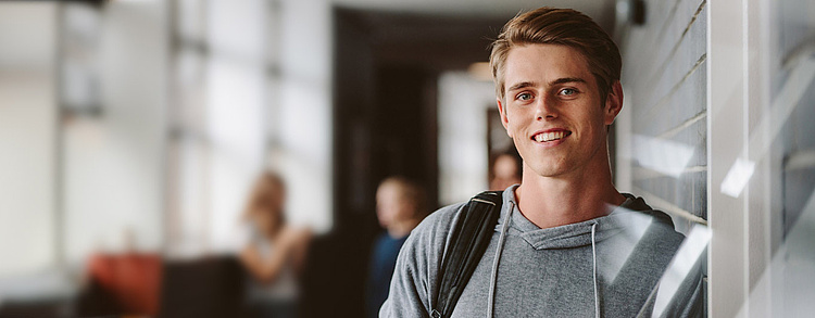 Ein junger Mann mit Rucksack lehnt lächelnd an einer Glaswand in einem hellen, modernen Gebäude. Im Hintergrund sind weitere Personen in einem unscharfen Bereich zu sehen.