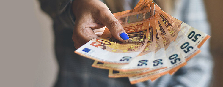 Eine Hand mit blau lackierten Fingernägeln hält mehrere 50-Euro-Scheine, vor einem unscharfen Hintergrund in neutralen Farben.