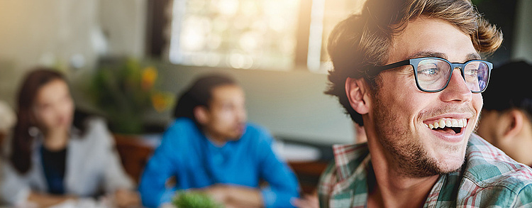 Ein junger Mann mit Brille und kariertem Hemd lacht und blickt freudig zur Seite. Im Hintergrund sind unscharf andere Menschen in einer hellen, freundlichen Umgebung zu sehen, die in ein Gespräch vertieft sind.
