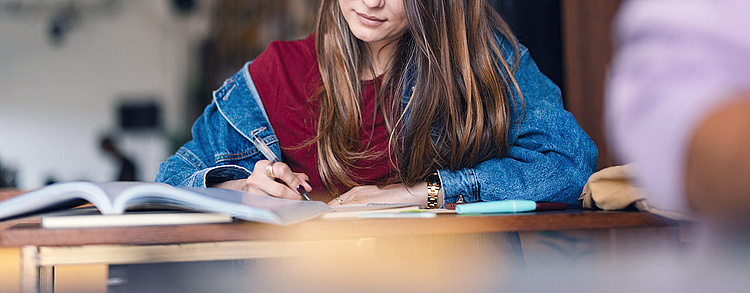 Eine junge Frau in einer Jeansjacke sitzt an einem Tisch und schreibt in ein Notizbuch. Vor ihr liegt ein aufgeschlagenes Buch, und im Hintergrund ist ein unscharfer Raum erkennbar.