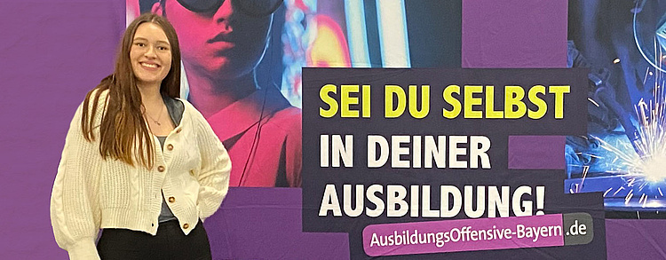 ine junge Frau steht lächelnd vor einer violetten Werbewand mit der Aufschrift „SEI DU SELBST IN DEINER AUSBILDUNG!“ und der Website „AusbildungsOffensive-Bayern.de“. Im Hintergrund sind stilisierte Bilder von Schweißen und urbanen Szenen zu sehen.