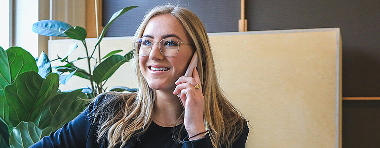 eine lächelnde Frau in einem dunkelblauen Blazer, die ein Telefonat führt und vor einem offenen silbernen MacBook sitzt. Sie sitzt auf einem beigen Sofa in einem modernen Büro oder Wohnzimmer mit Pflanzen im Hintergrund, was eine freundliche und professionelle Atmosphäre vermittelt.