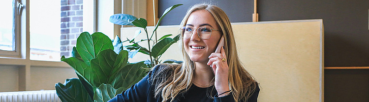 eine lächelnde Frau in einem dunkelblauen Blazer, die ein Telefonat führt und vor einem offenen silbernen MacBook sitzt. Sie sitzt auf einem beigen Sofa in einem modernen Büro oder Wohnzimmer mit Pflanzen im Hintergrund, was eine freundliche und professionelle Atmosphäre vermittelt.